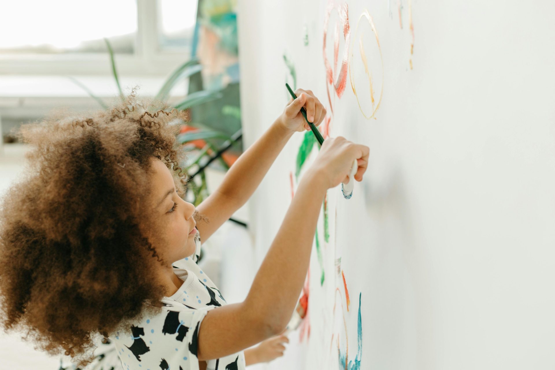 A young girl with curly hair paints creatively on a white wall in a bright indoor setting.