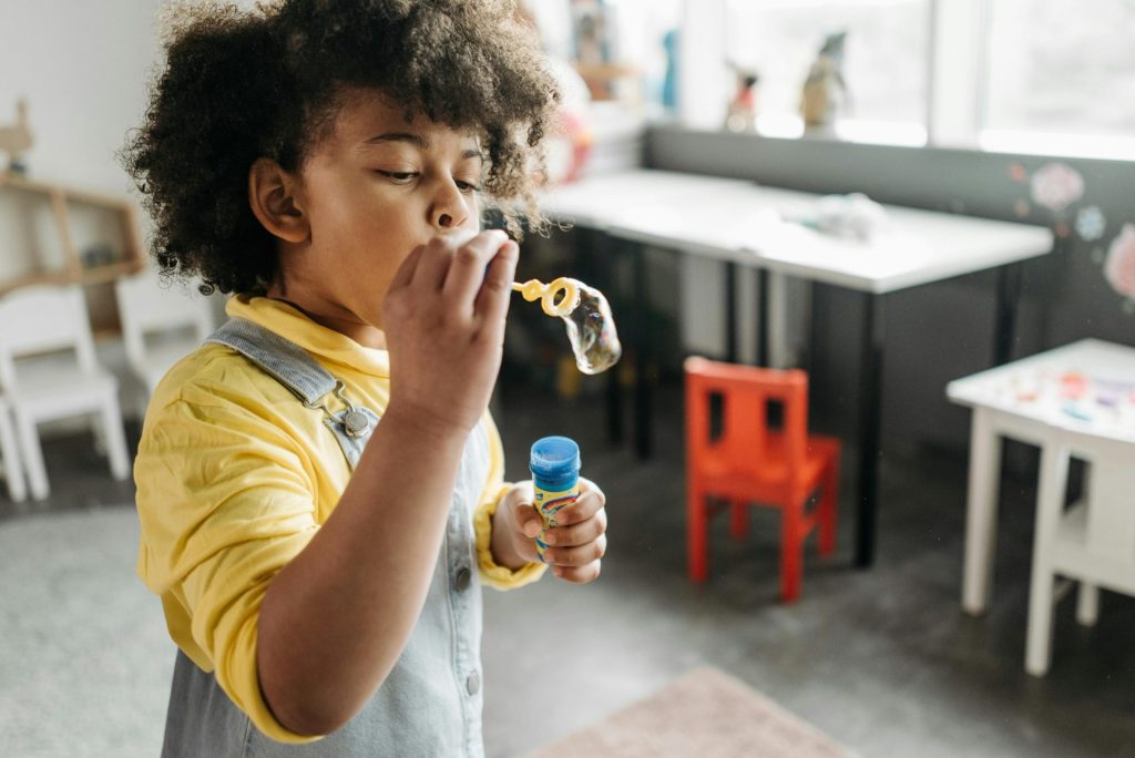 Cute child blowing bubbles in a playroom, wearing a yellow shirt and overalls.